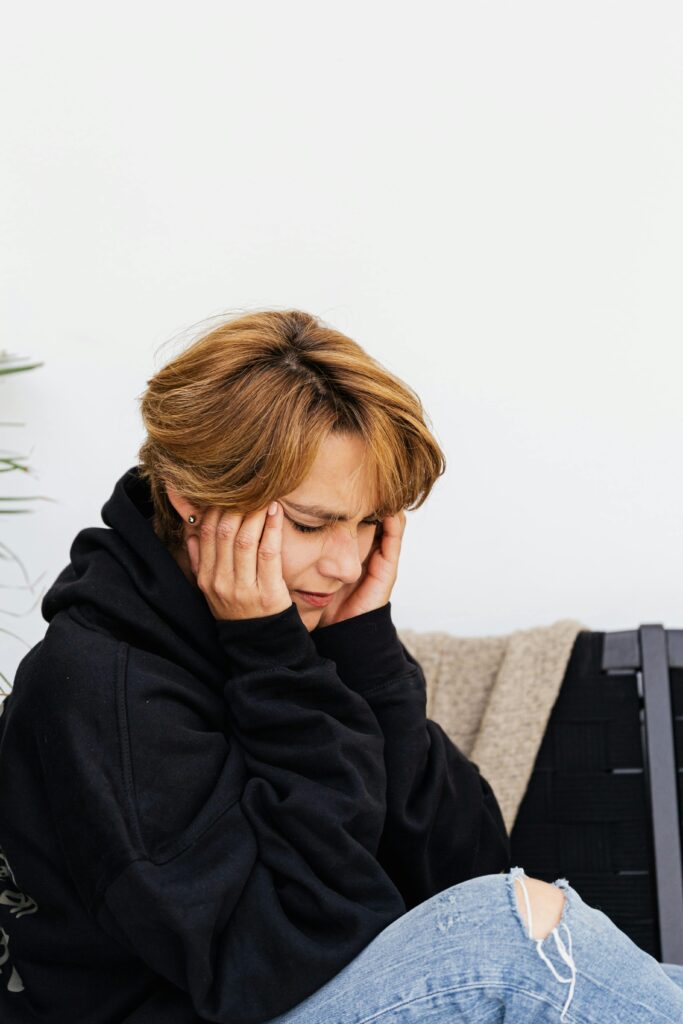A woman sitting on a couch, resting her hands on her head, appears deep in thought or contemplation.