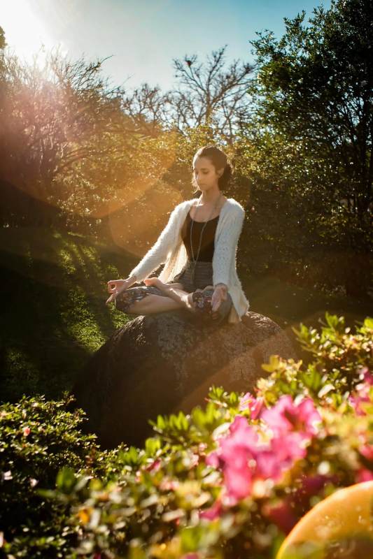 A woman sits cross-legged on a sunlit rock, meditating peacefully in a serene outdoor setting.