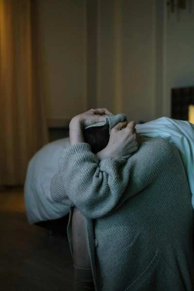 A woman sits on a bed in a dimly lit room, creating a somber and introspective atmosphere.
