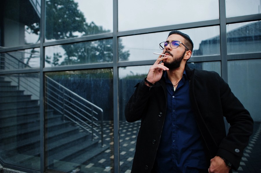 A man in a stylish suit smokes a cigarette, exuding a sophisticated and relaxed demeanor in a dimly lit setting.