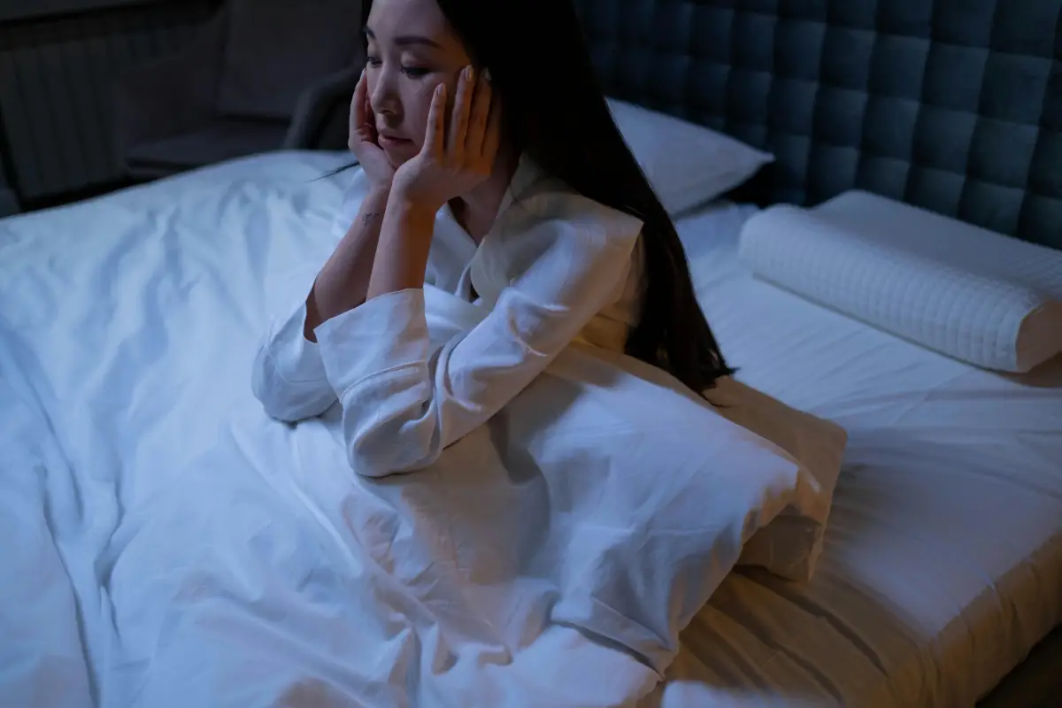 A woman sitting in bed, resting her hands on her face, appears contemplative or distressed.