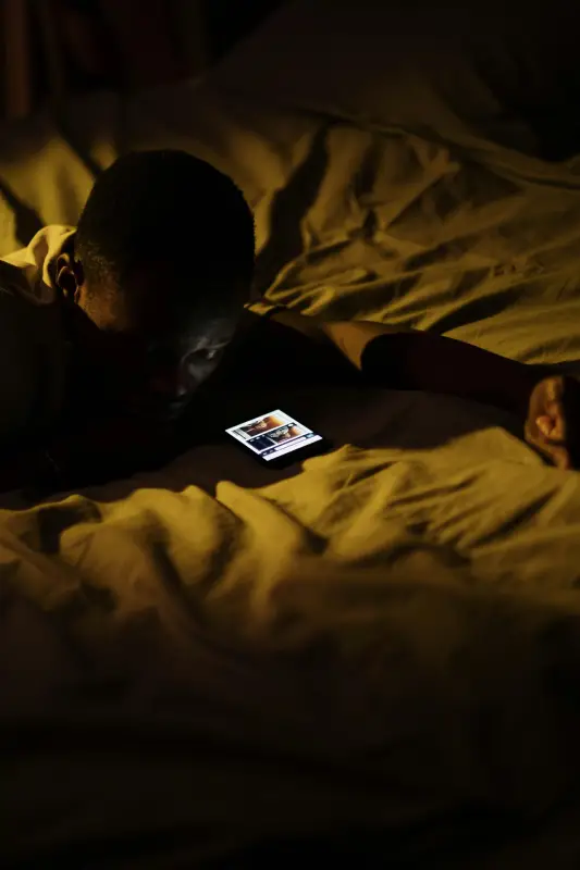 A young boy lying in bed, focused on his phone, with soft bedding and a cozy atmosphere surrounding him.