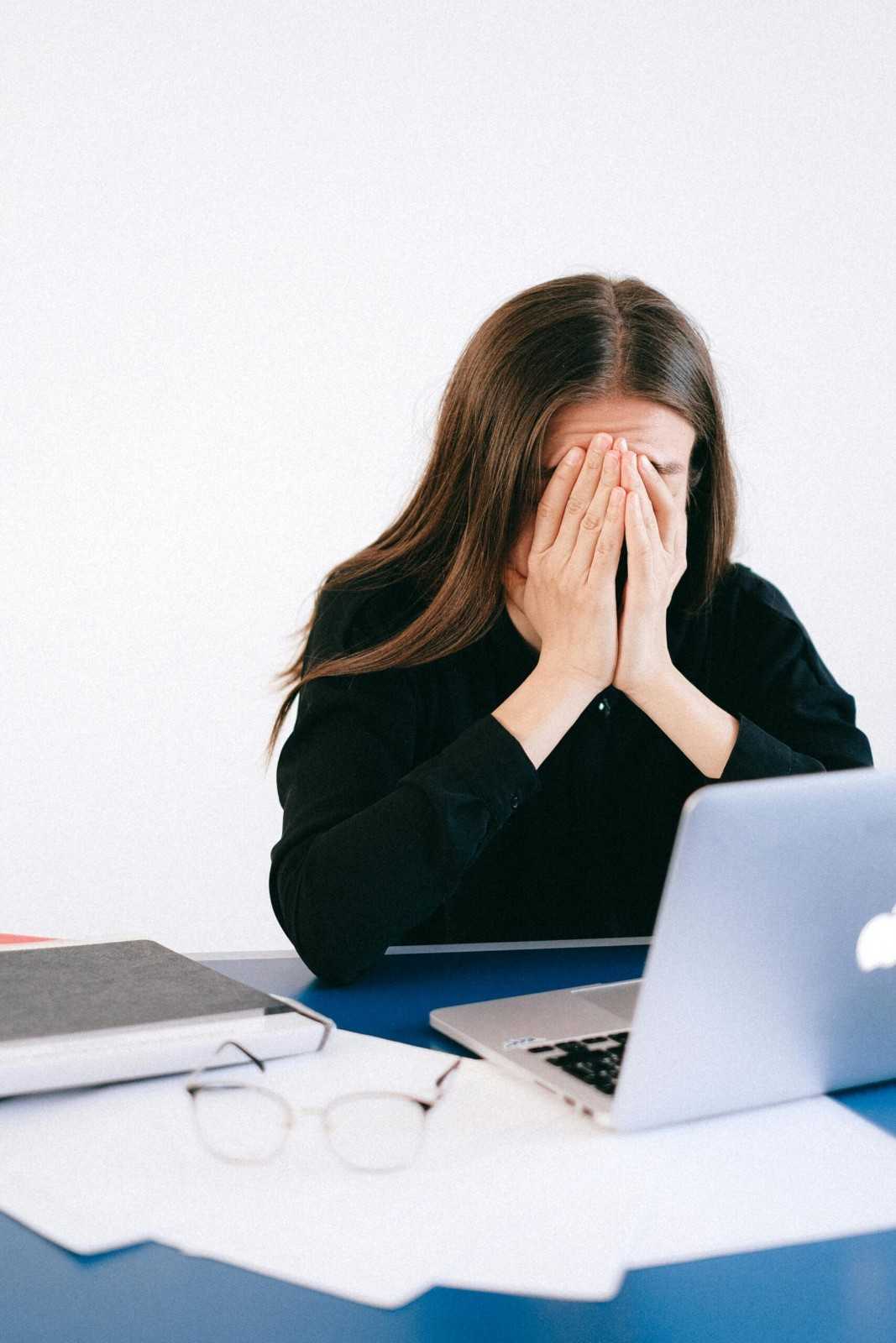 A woman sits at a desk, resting her face in her hands, appearing deep in thought or stressed.